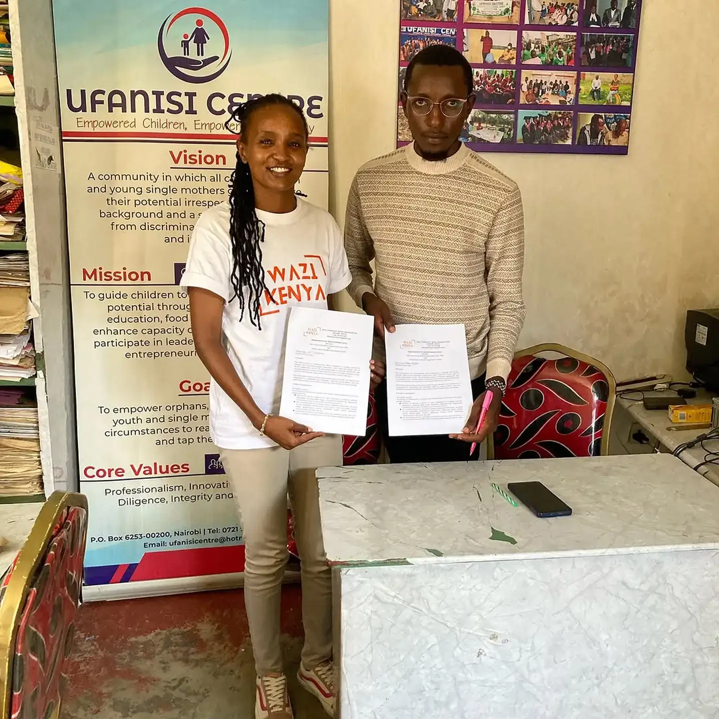 A Wazi Kenya volunteer in a white Wazi shirt and a colleague hold signed documents in front of a Ufanisi Centre banner.