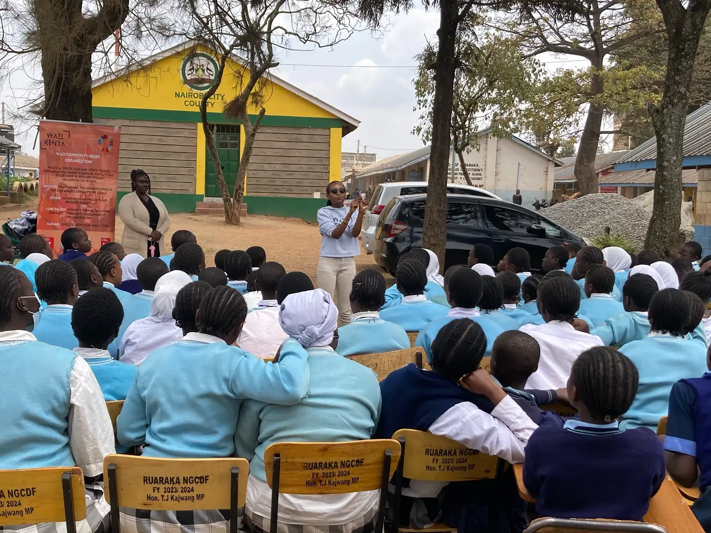A large outdoor school assembly fills rows of chairs beneath trees and a yellow classroom block.