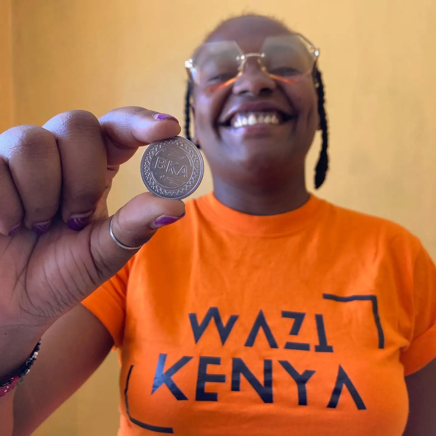 A smiling Wazi volunteer in an orange shirt holds a metal dispenser token close to the camera.