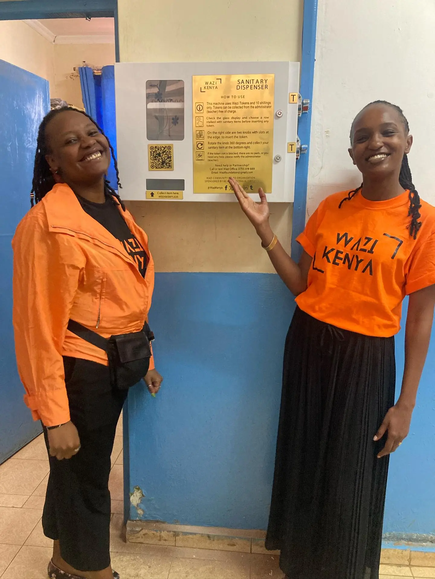 Two women in orange Wazi shirts smile beside a newly mounted dispenser, one gesturing toward it.