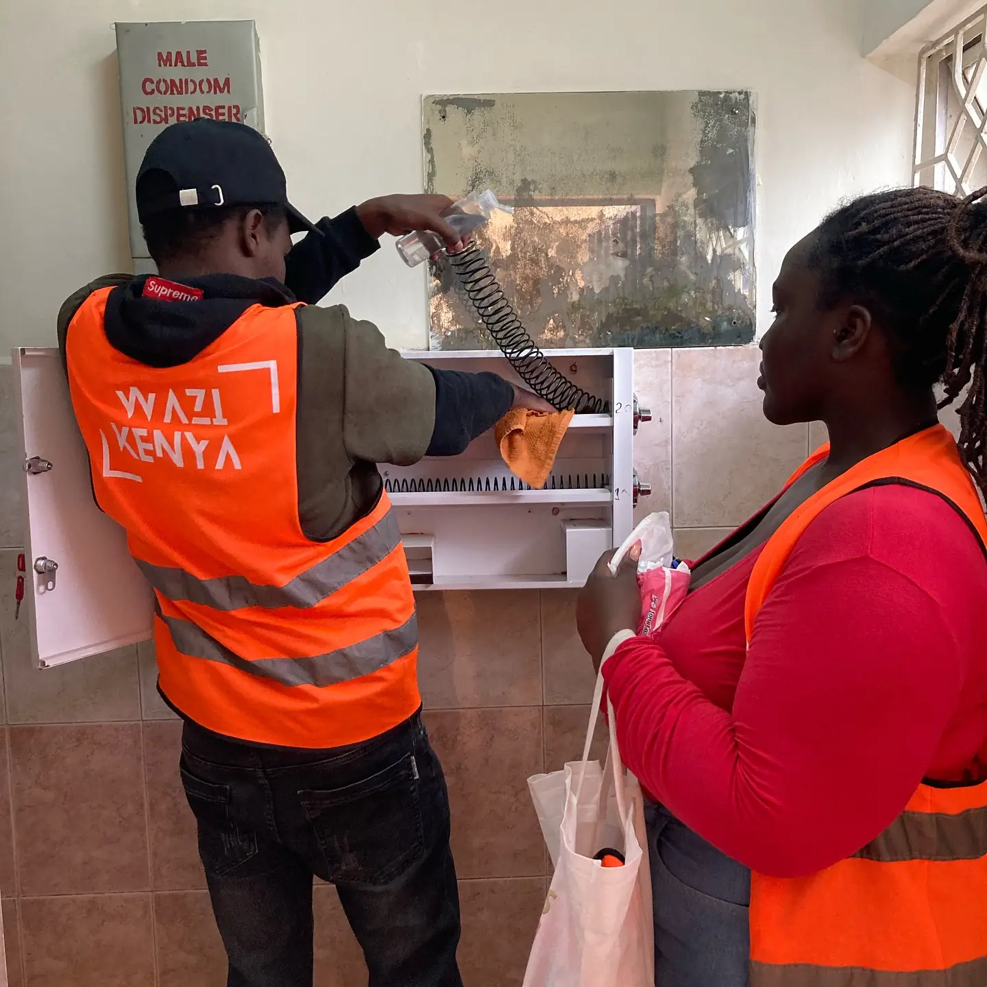 Two volunteers in orange Wazi vests work together at an open sanitary dispenser.