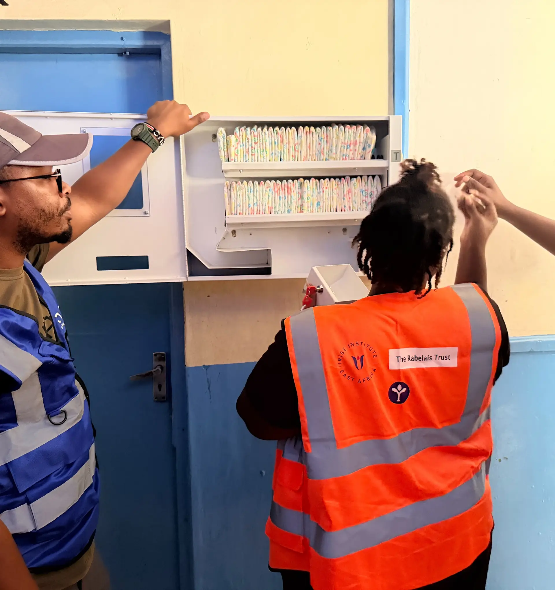 Wazi and CAEL volunteers open a stocked wall dispenser in a blue and yellow corridor.