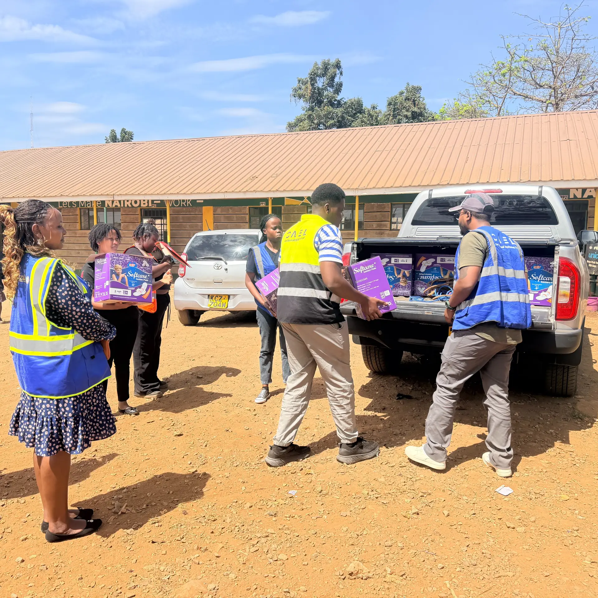 Partners carry pad cartons from a pickup across a sunlit school compound.