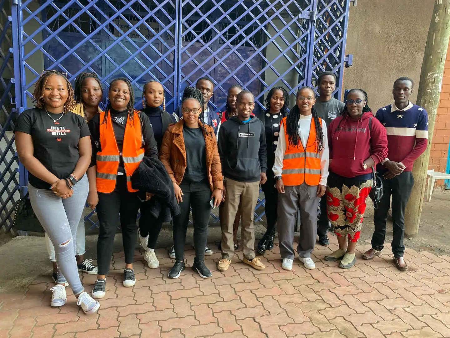 Wazi volunteers gather shoulder to shoulder on brick paving outside a blue lattice doorway.