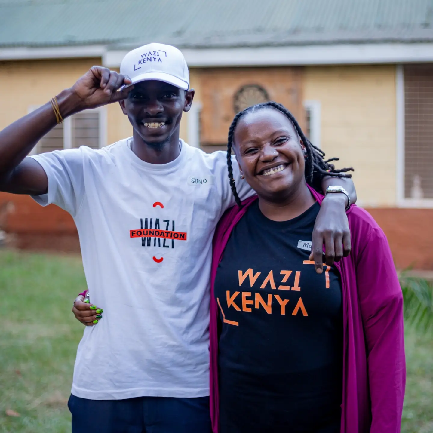 Two volunteers stand arm in arm laughing, one lifting a white Wazi cap.