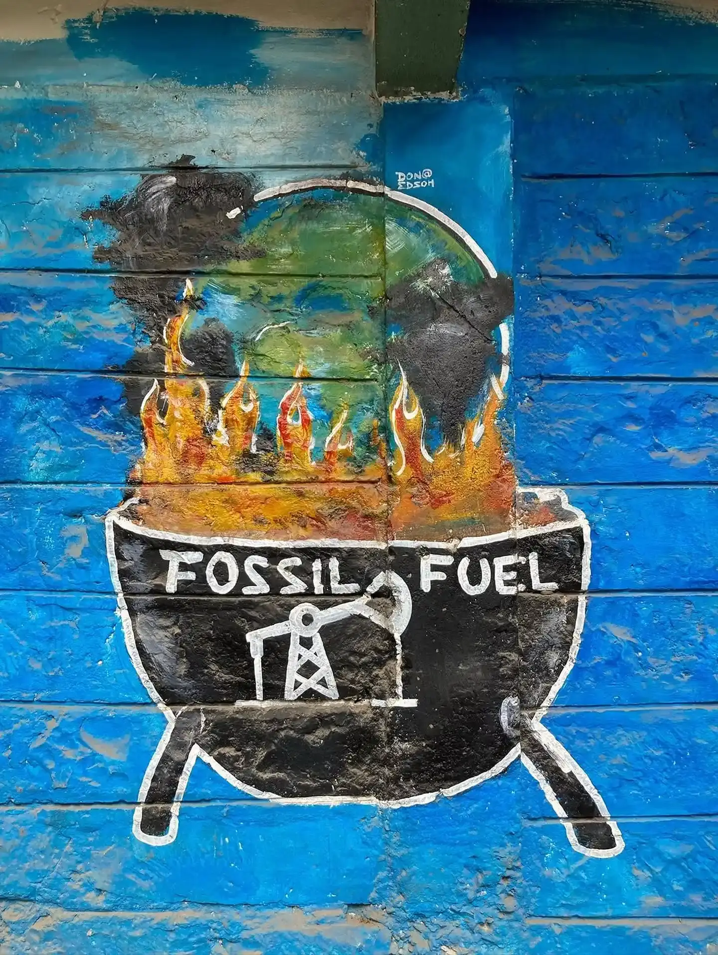 A painted wall mural of the Earth burning in a cauldron labelled "Fossil Fuel", on a bright blue wall.