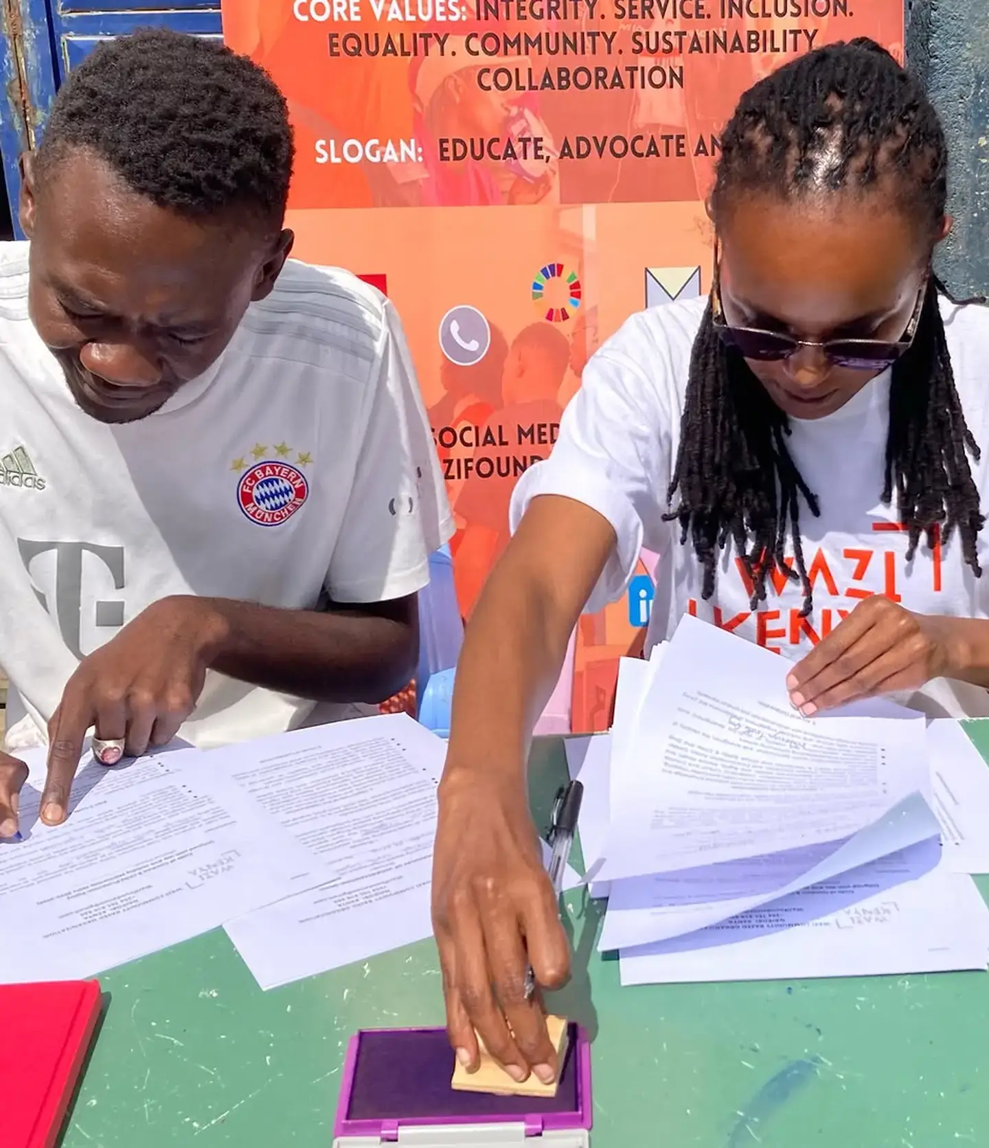 Two adults in Wazi Kenya shirts sit at a table, one stamping and the other turning through signed paperwork.
