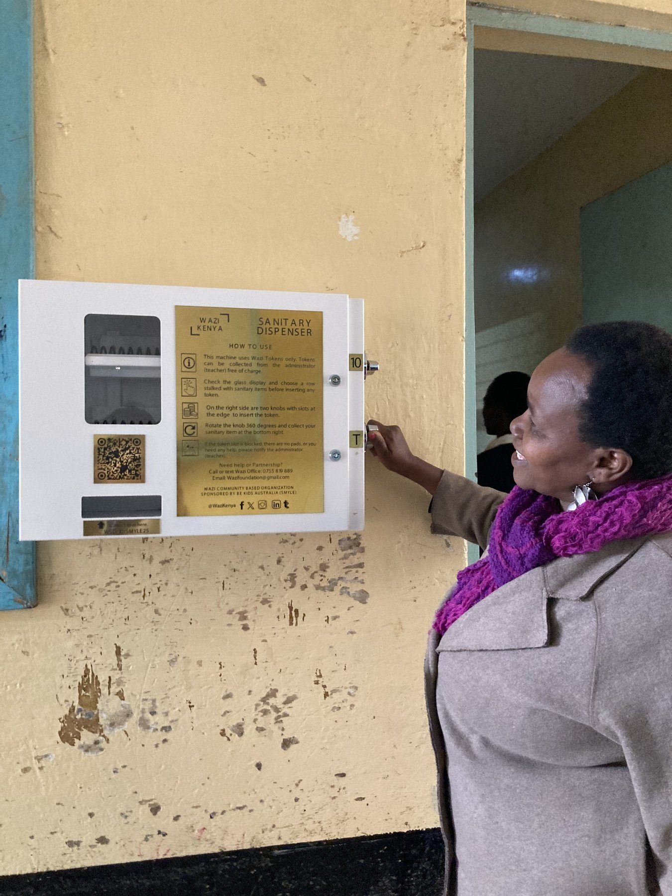 A Wazi sanitary dispenser mounted on a school wall with the gold instruction label and QR code visible.